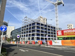 An urban construction site features a partially built multi-story office building surrounded by fencing and signs promoting the development. Several cranes are visible, suggesting ongoing construction. Various modern buildings and skyscrapers are in the background under a clear blue sky with light cloud streaks.