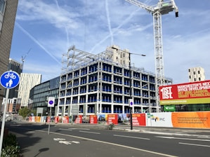 An urban construction site features a partially built multi-story office building surrounded by fencing and signs promoting the development. Several cranes are visible, suggesting ongoing construction. Various modern buildings and skyscrapers are in the background under a clear blue sky with light cloud streaks.