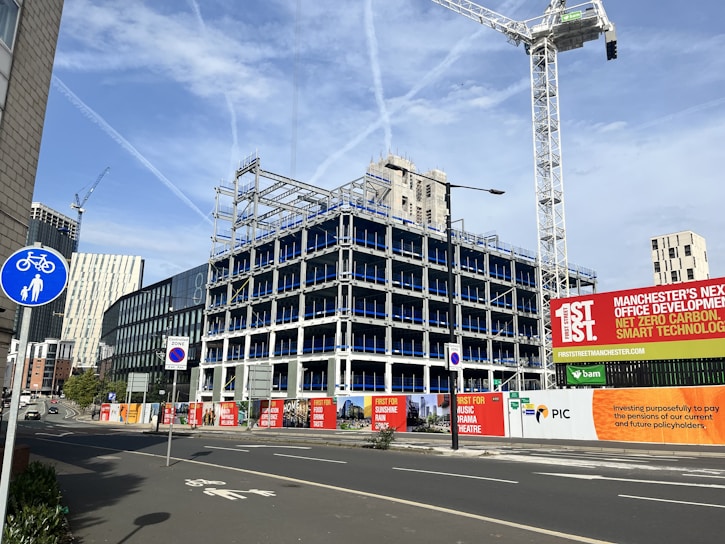An urban construction site features a partially built multi-story office building surrounded by fencing and signs promoting the development. Several cranes are visible, suggesting ongoing construction. Various modern buildings and skyscrapers are in the background under a clear blue sky with light cloud streaks.