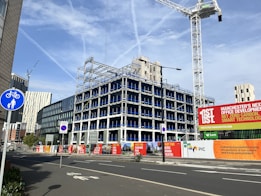 An urban construction site features a partially built multi-story office building surrounded by fencing and signs promoting the development. Several cranes are visible, suggesting ongoing construction. Various modern buildings and skyscrapers are in the background under a clear blue sky with light cloud streaks.