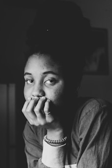 A black and white portrait of a person gazing forward with a contemplative expression. They are resting their chin on their hand, wearing a sweater and a beaded bracelet. The lighting creates shadows on their face, enhancing the introspective mood.