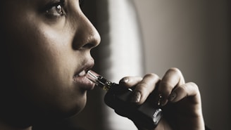 A person is holding a vape device close to their lips, with a contemplative expression. The lighting is dim and dramatic, focusing on their face and hand.