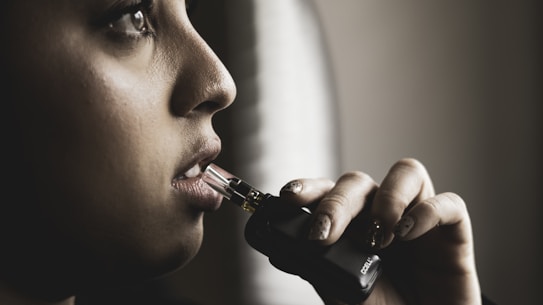 A person is holding a vape device close to their lips, with a contemplative expression. The lighting is dim and dramatic, focusing on their face and hand.