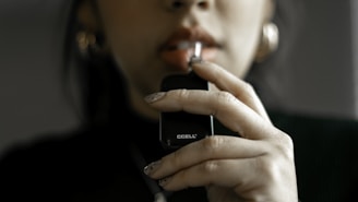 A person is holding a black vaping device close to their lips. They have neatly manicured nails with a neutral design, and are wearing large hoop earrings.