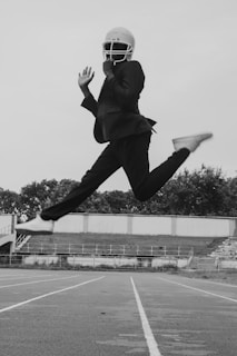 A person wearing a suit and a helmet is captured mid-air, leaping across a running track. The background features empty bleachers and a line of trees.