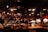 A warm, dimly lit restaurant interior with wooden tables and chairs, where several people are seated and engaged in conversation or dining. The atmosphere is enhanced by hanging industrial-style light fixtures and various decorations on the walls. The setting feels cozy and inviting.