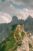 A narrow hiking trail winds along a grassy ridge, with a backdrop of jagged mountain peaks under a partly cloudy sky. Several hikers are visible on the path, enjoying the scenic cliffs.