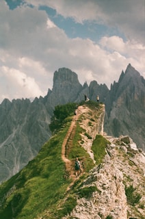 Travelers crossing a narrow mountain path with panoramic views of rocky cliffs and blue skies.