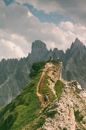 A narrow hiking trail winds along a grassy ridge, with a backdrop of jagged mountain peaks under a partly cloudy sky. Several hikers are visible on the path, enjoying the scenic cliffs.