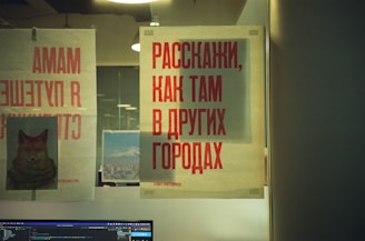 Various posters are displayed on a glass wall. One prominently features the text in red Cyrillic letters, another depicts an image of a fox with a cozy scarf, and a third shows a landscape photograph of a city with mountains in the background. Below, a computer monitor displays lines of code against a dark background. Overhead lights create reflections on the glass.