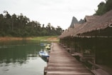 A wooden walkway extends alongside a series of thatched-roof huts beside a tranquil body of water. The surrounding scenery includes lush green forests and distant mountains under a cloudy sky. Several kayaks in various colors are moored beside the walkway, adding a hint of activity to the peaceful setting.