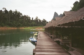 A wooden walkway extends alongside a series of thatched-roof huts beside a tranquil body of water. The surrounding scenery includes lush green forests and distant mountains under a cloudy sky. Several kayaks in various colors are moored beside the walkway, adding a hint of activity to the peaceful setting.