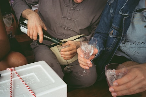 A group of people gathered around, one person pouring a drink into a glass filled with ice cubes. The individuals appear to be casually dressed, with attention focused on the pouring action. A white cooler with red rope detailing is visible in the foreground.