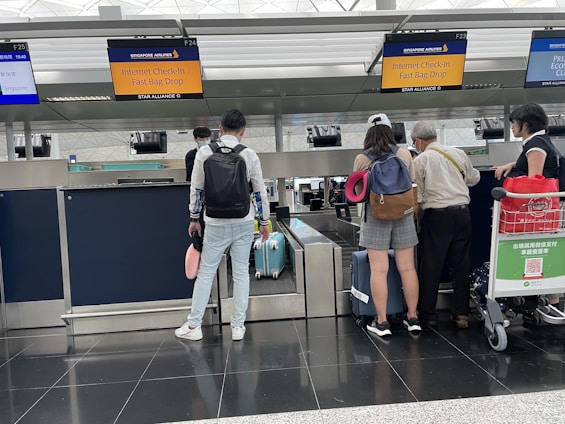 A group of travelers stand at an airport check-in counter, with visible signage for Singapore Airlines. They are waiting with their luggage, which includes suitcases and travel bags. One person is carrying a neck pillow, and another has a red trolley containing a QR code for payment. The area is well-lit, with check-in screens and information displayed overhead.