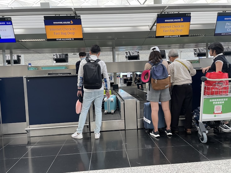A group of travelers stand at an airport check-in counter, with visible signage for Singapore Airlines. They are waiting with their luggage, which includes suitcases and travel bags. One person is carrying a neck pillow, and another has a red trolley containing a QR code for payment. The area is well-lit, with check-in screens and information displayed overhead.