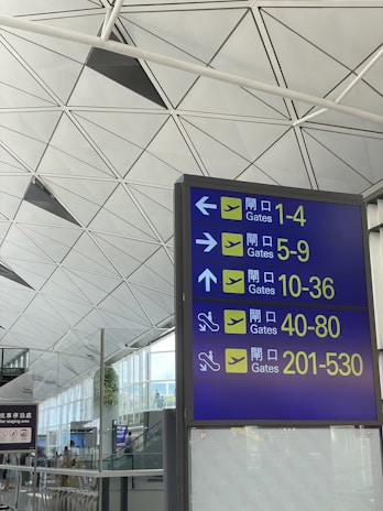 A large airport sign is displayed, directing passengers to various departure gates, numbered 1-530, using arrows and icons. The background features a modern ceiling design with geometric shapes and a glimpse of a spacious airport interior.