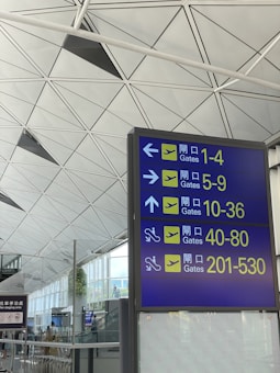 A large airport sign is displayed, directing passengers to various departure gates, numbered 1-530, using arrows and icons. The background features a modern ceiling design with geometric shapes and a glimpse of a spacious airport interior.