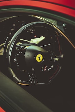 A close-up view of a car's steering wheel featuring a luxury brand logo at its center. The interior is sleek and designed with high-quality materials, including carbon fiber accents and a digital dashboard in the background.