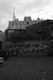 There is a wall with graffiti art that says 'Under Construction' in large, stylish letters. The wall is part of a background scene with older buildings, one of which has a classical, ornate design with multiple circular windows and a white spire. The sky looks overcast, and there are a few cars parked in the foreground on a gravel surface.