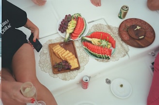 A casual setting with a variety of snacks arranged on decorative plates. There are artfully sliced watermelons garnished with pineapples, a plate with crackers and grapes, and another with slices of salami and cheese. Surrounding the food are several beverages, including a can of soda and a can of beer, and the bottom of a glass held in someone's hand is visible.