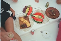 A casual setting with a variety of snacks arranged on decorative plates. There are artfully sliced watermelons garnished with pineapples, a plate with crackers and grapes, and another with slices of salami and cheese. Surrounding the food are several beverages, including a can of soda and a can of beer, and the bottom of a glass held in someone's hand is visible.
