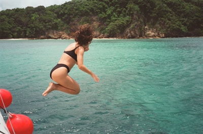 A person wearing a black swimsuit is caught in mid-air as they jump into crystal clear turquoise water from a boat. There are red buoys attached to the boat close to the edge of the frame. A lush green island with rocky shoreline is in the background under an overcast sky.