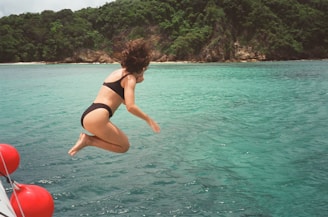 A person wearing a black swimsuit is caught in mid-air as they jump into crystal clear turquoise water from a boat. There are red buoys attached to the boat close to the edge of the frame. A lush green island with rocky shoreline is in the background under an overcast sky.