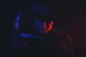 A sinister figure is partially illuminated by a dramatic play of red and blue lighting. The figure wears a dark hat and has a menacing expression, evoking a sense of fear and mystery.