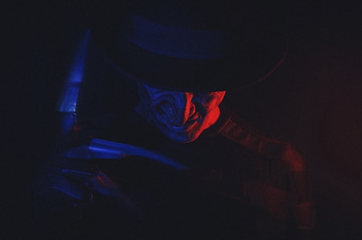 A sinister figure is partially illuminated by a dramatic play of red and blue lighting. The figure wears a dark hat and has a menacing expression, evoking a sense of fear and mystery.