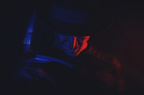 A sinister figure is partially illuminated by a dramatic play of red and blue lighting. The figure wears a dark hat and has a menacing expression, evoking a sense of fear and mystery.