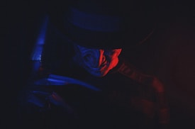 A sinister figure is partially illuminated by a dramatic play of red and blue lighting. The figure wears a dark hat and has a menacing expression, evoking a sense of fear and mystery.