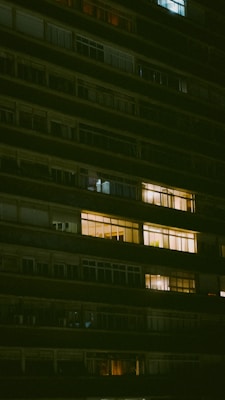 A high-rise building facade at night with various illuminated windows, some brightly lit creating a warm atmosphere, while others remain dark. The building appears residential with multiple stories and a mix of different window styles.