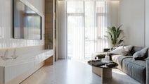 A modern living room with a large flat-screen TV mounted on the wall, a sleek white media console below, and a comfortable gray sectional sofa adorned with cushions. A round coffee table with books and a candle sits in front of the sofa. Floor-to-ceiling windows allow natural light to brighten the space, and a large potted plant adds a touch of greenery. The room features a neutral color palette with elegant decor.