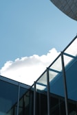 Modern architectural structure with glass windows and metal frames, set against a bright blue sky with scattered clouds.