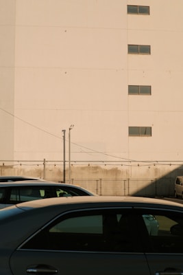 A large beige building wall with small windows at the top is situated behind a parking lot with a few parked cars. The shadow of the building partially covers the parking lot. A security camera and wire fencing are present, adding to the urban setting.