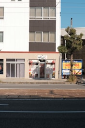 A street view of a building with a small storefront on the ground floor. The facade has a combination of white and dark gray panels with several windows covered by beige curtains. The storefront has a sign in Japanese, flanked by decorative red lanterns. A tree is situated to the right side on the sidewalk, and a blue sign with pricing information is visible nearby. The scene includes a paved road in the foreground.