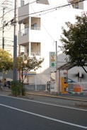 A street scene featuring a parking area with a payment kiosk and a parked car. The area is bordered by a low fence and there are several trees nearby. A multi-story building with balconies is visible in the background, and overhead utility lines run across the scene.