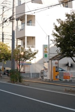A street scene featuring a parking area with a payment kiosk and a parked car. The area is bordered by a low fence and there are several trees nearby. A multi-story building with balconies is visible in the background, and overhead utility lines run across the scene.