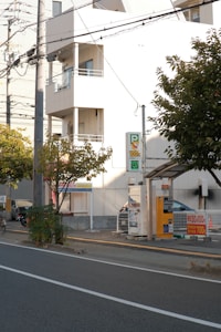 A street scene featuring a parking area with a payment kiosk and a parked car. The area is bordered by a low fence and there are several trees nearby. A multi-story building with balconies is visible in the background, and overhead utility lines run across the scene.