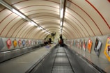 A long, steep escalator in an underground subway station with a curved, tiled ceiling. The walls are lined with colorful advertisements. Several people are riding the escalator, and a few are standing at the top. The lighting is fluorescent, creating a utilitarian atmosphere.