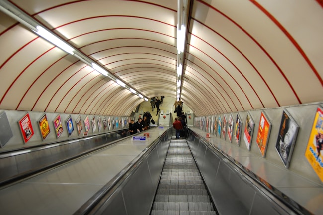 A long, steep escalator in an underground subway station with a curved, tiled ceiling. The walls are lined with colorful advertisements. Several people are riding the escalator, and a few are standing at the top. The lighting is fluorescent, creating a utilitarian atmosphere.