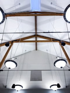 A modern ceiling design featuring wooden beams and multiple circular light fixtures. The ceiling has a high pitch with a skylight at the top center, and the beams form a triangular pattern. Several electrical wires and cables are arranged in a neat and organized manner.