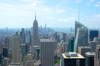 A bustling urban skyline featuring an array of skyscrapers under a bright blue sky. The iconic Empire State Building stands prominently among other tall buildings. In the distance, the waters sparkle, separating parts of the city landscape.