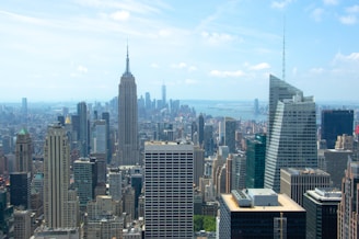 A bustling urban skyline featuring an array of skyscrapers under a bright blue sky. The iconic Empire State Building stands prominently among other tall buildings. In the distance, the waters sparkle, separating parts of the city landscape.