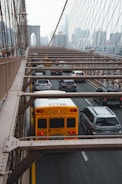 A yellow school bus and several cars are driving on a busy road with metal beams overhead. The road is part of a suspension bridge leading toward a city skyline visible in the background.