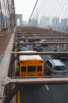 A yellow school bus and several cars are driving on a busy road with metal beams overhead. The road is part of a suspension bridge leading toward a city skyline visible in the background.