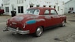 A vintage red car with visible wear and rust is parked on a cracked pavement in front of a weathered, white building. The car has a 'for sale' sign in the window, and its paint shows signs of age with patches of discoloration. The area appears to be an old, possibly abandoned service station with scattered debris and a feeling of neglect.