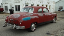 A vintage red car with visible wear and rust is parked on a cracked pavement in front of a weathered, white building. The car has a 'for sale' sign in the window, and its paint shows signs of age with patches of discoloration. The area appears to be an old, possibly abandoned service station with scattered debris and a feeling of neglect.