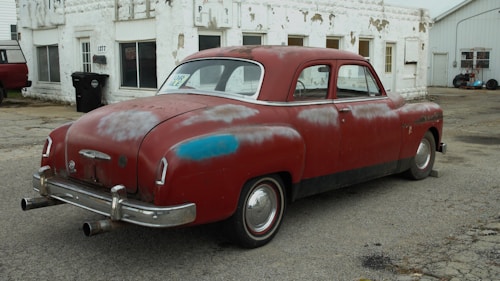 A vintage red car with visible wear and rust is parked on a cracked pavement in front of a weathered, white building. The car has a 'for sale' sign in the window, and its paint shows signs of age with patches of discoloration. The area appears to be an old, possibly abandoned service station with scattered debris and a feeling of neglect.
