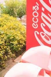 A vibrant outdoor scene with a large red sign featuring white lettering partially visible. Yellow flowers and lush green foliage occupy the left side of the image, and a paved path is visible at the bottom.